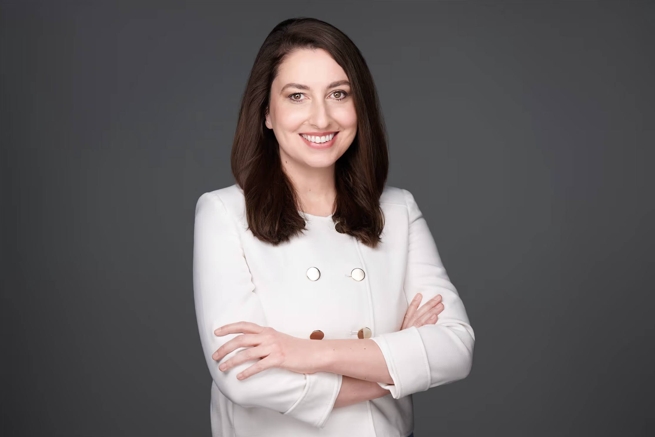 Smiling woman with long brown hair wearing a white blazer, standing with arms crossed against a gray background.
